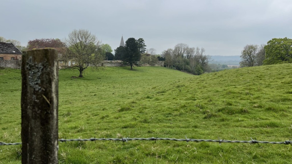 View of a green open field with a wooden fence post in the foreground and a wire fence running through the image. In the background, trees surround a historic stone wall, with a church steeple rising above. The scene is typical of rural England, offering potential land for development, making it relevant for discussions about the call for sites process in planning and land promotion.