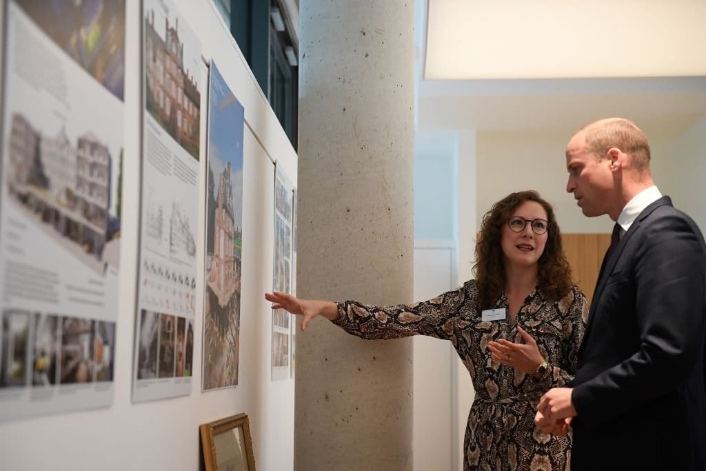 Professional woman presenting architectural project displays to William, Prince of Wales, discussing urban building designs in a gallery setting with visible posters on the wall.
