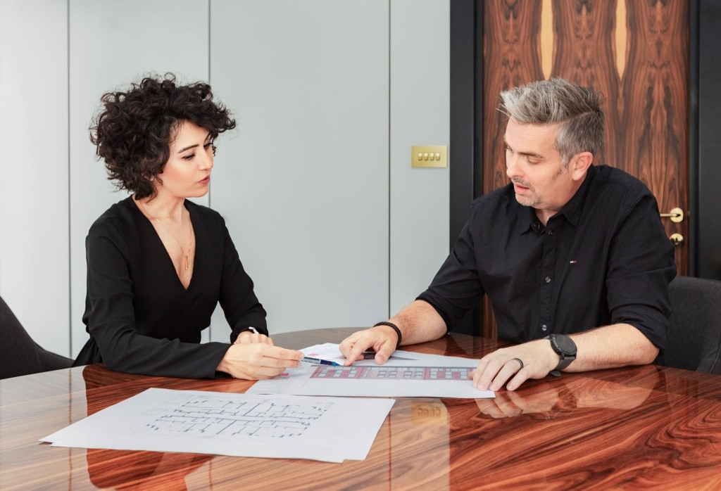 Two RIBA chartered architects in professional attire engaged in a focused discussion over architectural plans on a polished wooden table in a modern office environment.