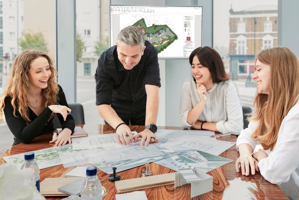 Enthusiastic team of architects laughing and collaborating around a table with building plans, architectural models, and a 3D site plan on a computer monitor in a bright office with a London city view.