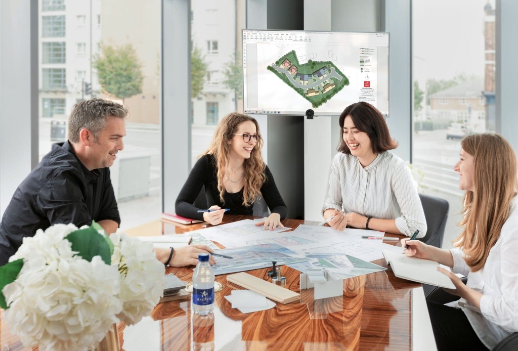 Group of four smiling architects discussing plans around a table with a 3D building model displayed on a screen in a modern, light-filled office with city views in the background.