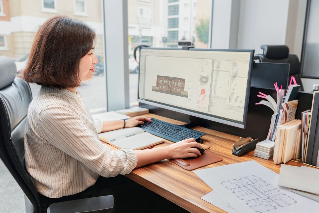 Female architect working on architectural software at her workstation with multiple building plans on the screen, in a modern office environment with books and documents on the desk.