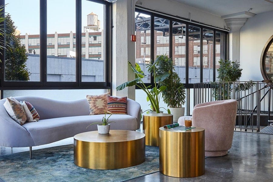 Elegant urban lounge area with a modern curved sofa, brass coffee tables, chic accent pillows, and indoor plants, complemented by large windows with a cityscape view.