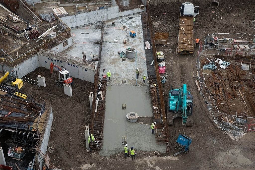Overhead view of construction workers and machinery at a redevelopment site, formerly a petrol station, now being transformed into residential housing. The image captures the foundation laying and structural work, showcasing the extensive process of converting disused petrol stations into valuable housing developments.