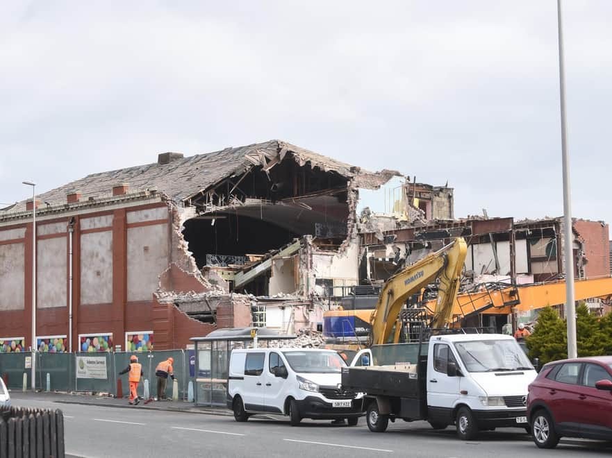 Street view of a large red-bricked triple storey bingo hall demolition of buildings planning permission with yellow bulldozers and contractors in orange hi-vis and hard hats in front of the hoarding.