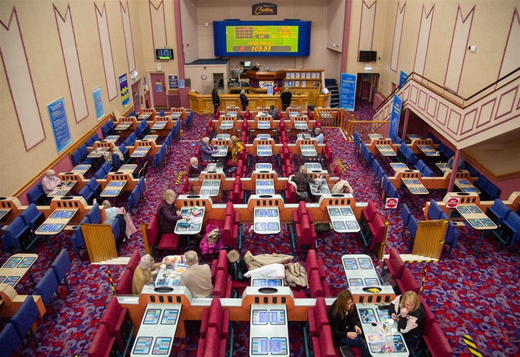 Downward view from the ceiling height of the interior of a large bingo hall with bright pink and blue foldable chairs around each table of 4 with couples and singles betting on the games