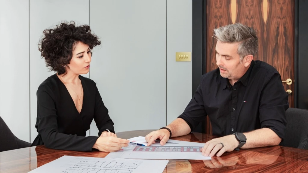 Nicole I. Guler, chartered town planner, and Robin Callister, chartered architect, reviewing floor plans and discussing an amalgamation project for residential properties in Belgravia, London. They are seated at a polished wooden table at Urbanist Architecture's office, analysing layouts and planning strategies.