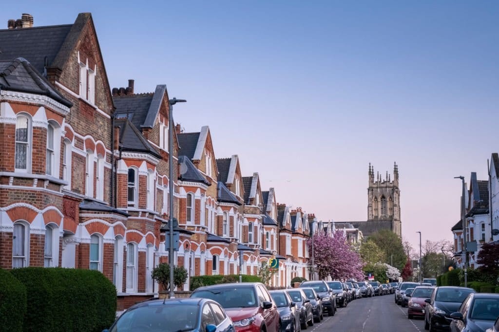 Row of charming Victorian houses with ornate red brick facades and white trim lining a quiet street, with blossoming trees and a historic church tower in the background at dusk.
