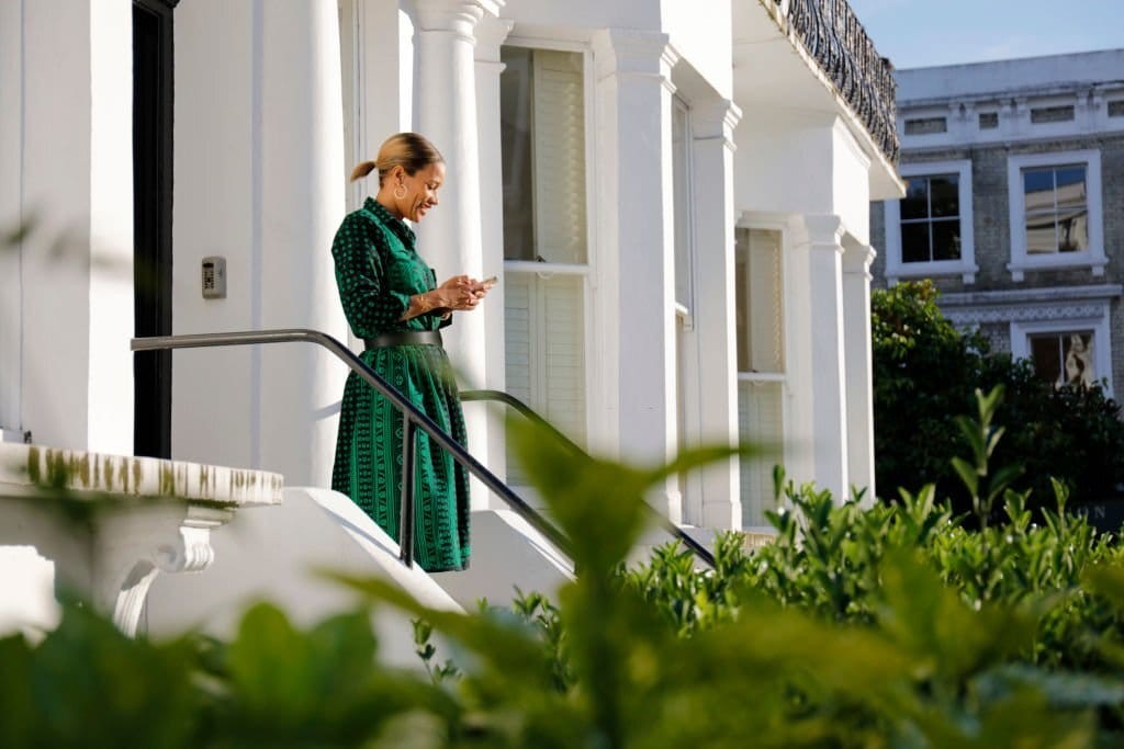 Elegant woman in a green dress smiling while texting on her phone, standing on the steps of a classic white-columned London townhouse with lush greenery in the foreground.
