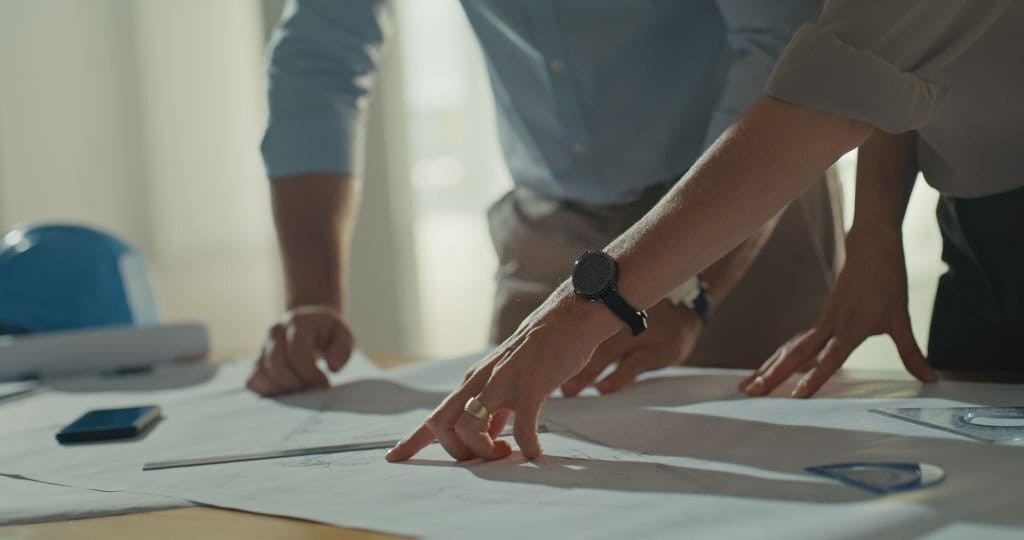 Close-up of professionals collaborating over architectural plans on a table, with focus on their hands pointing at a blueprint, a hardhat and smartphone in the background indicating a construction project discussion.