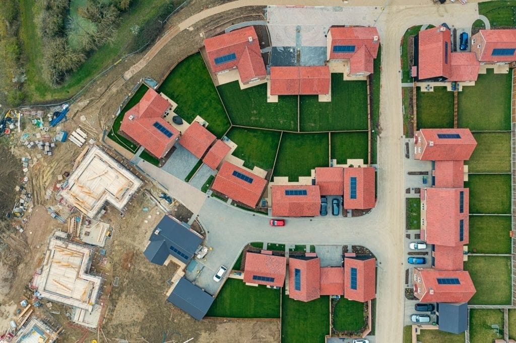 Aerial view of a new housing development under construction, with several completed houses with red roofs, new foundations in progress, and construction materials scattered on site, showcasing different stages of building and planning.