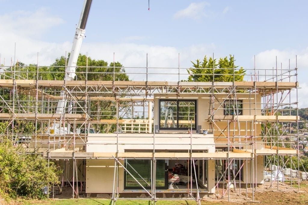 Mid-construction phase of a modern residential home with exposed wooden framing and extensive scaffolding, featuring a crane in operation, set against a backdrop of greenery and a clear sky, symbolising new home construction.