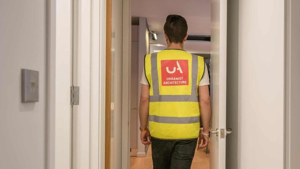 Construction professional from Urbanist Architecture wearing a high-visibility vest with a company logo, walking through a modern interior space during an on-site project inspection.