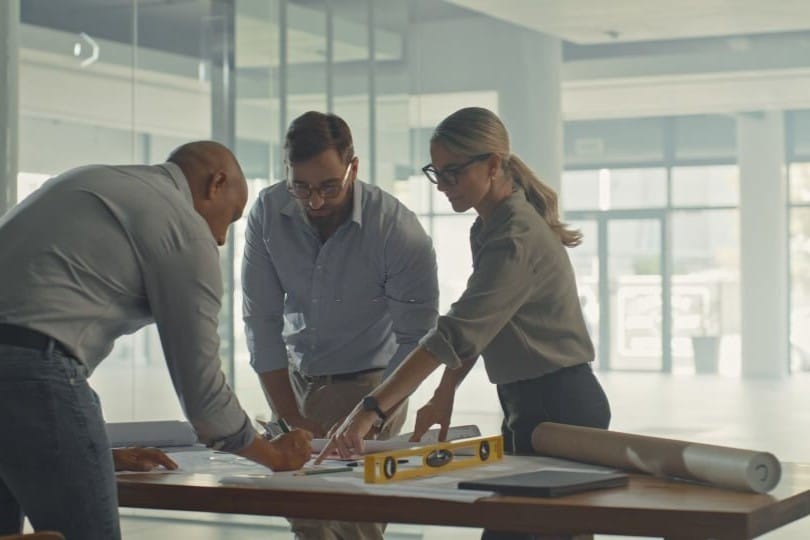 Two male architects in a grey and blue shirt discussing around a wooden table and marking with pens on the construction drawings with another female architect with glasses and a ponytail in the middle of an empty large commercial unit