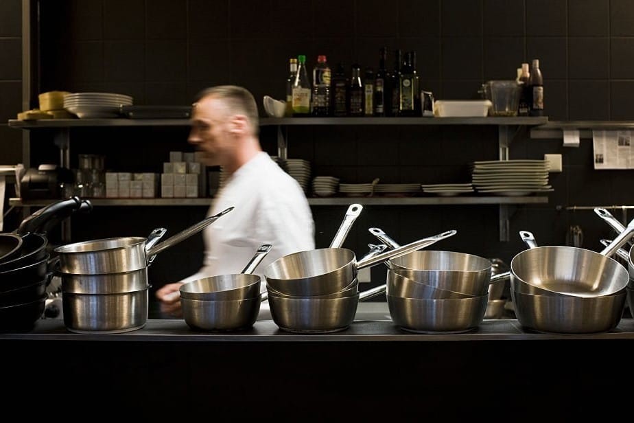 Moving and blurry chef in a white blouse and in a narrow and wide kitchen, walking behind a countertop displaying 6 stacks of stainless steel pots