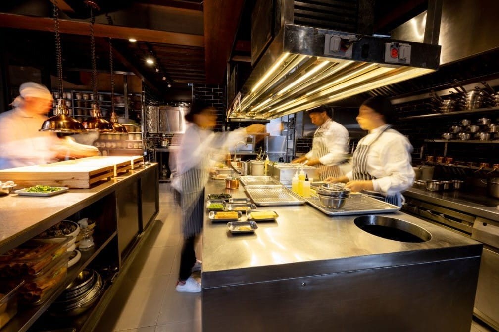 Long exposure photograph of four blurred chefs in white blouses and striped aprons working around two stainless steel kitchen islands with food heat lamps and ventilators