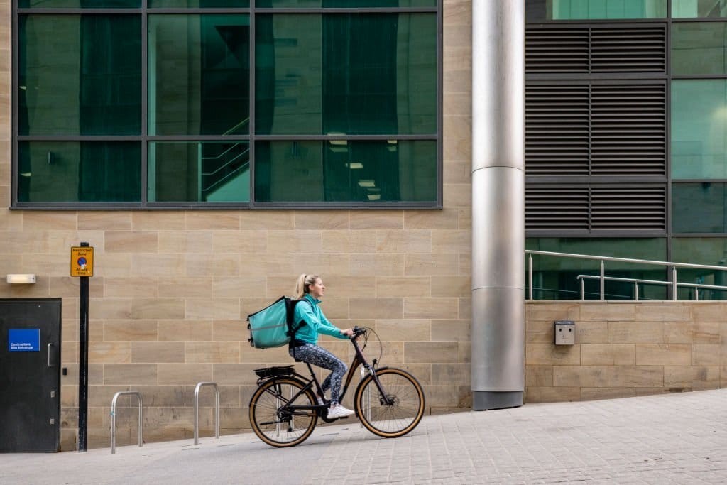 Blonde female dressed in the blue Deliveroo sweatshirt and bag riding her black and yellow tire bicycle in front of a large residential development with tall reflective windows