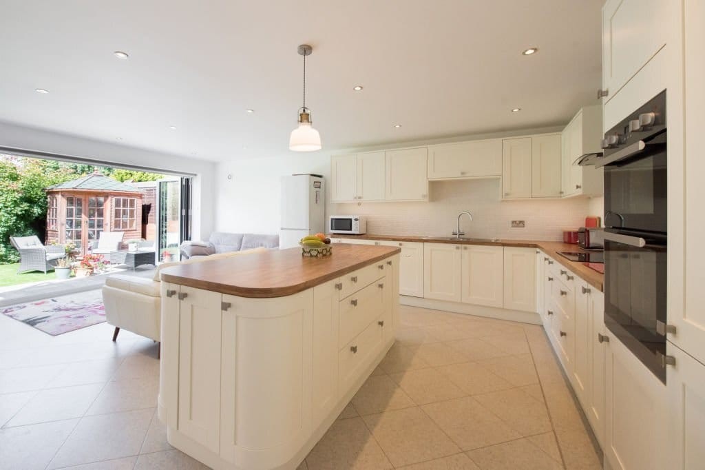 Modern kitchen rear extension in London with white walls, white cabinets and natural wood countertop with bi-fold doors leading to a lush green and sunny garden