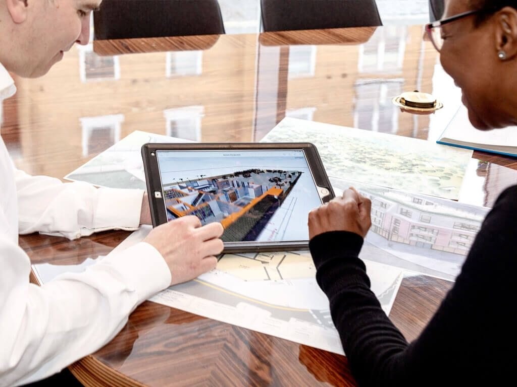 Two architects reviewing a detailed 3D building model on a tablet during a planning session. The image captures a collaborative design process, with blueprints and architectural drawings spread out on the polished wooden table. 