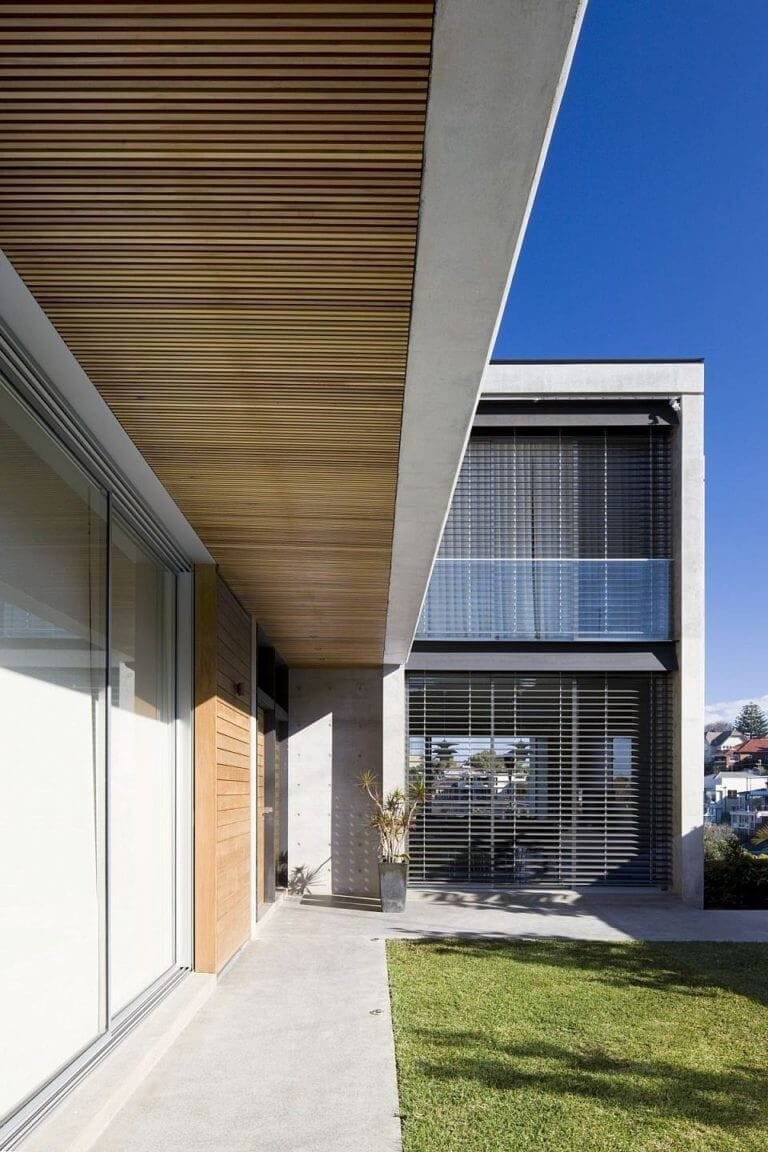 Contemporary architecture featuring a mix of concrete, wood, and glass materials with a stylish wooden ceiling overhang, large windows, and a second-story balcony with privacy screens.