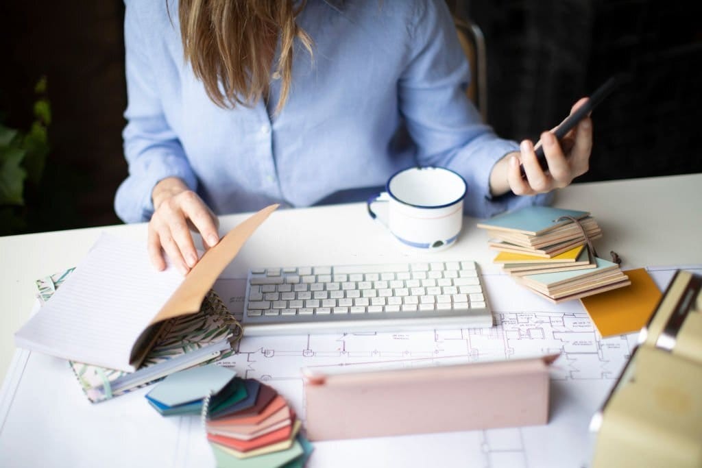 Interior designer planning a home renovation, with architectural blueprints, color swatches, and a keyboard on the desk, while using a smartphone, embodying the creative process in design.