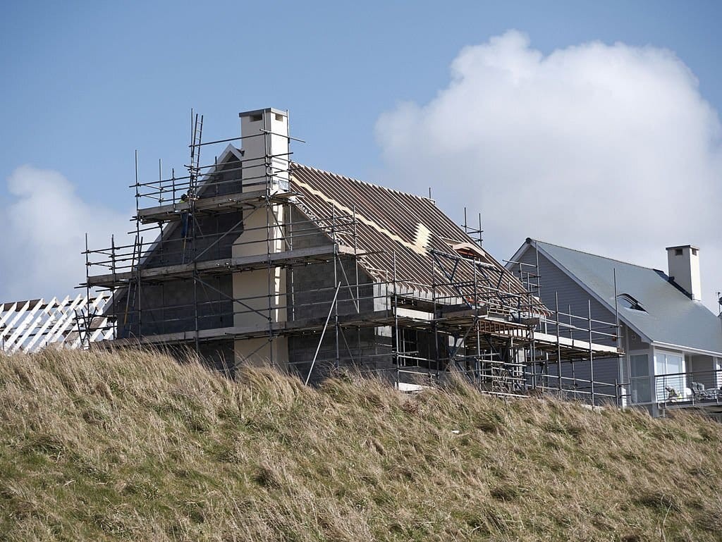 Renovation in progress on a coastal home, with scaffolding surrounding the structure as a new roof is being installed, set against a clear blue sky on a grassy hill.