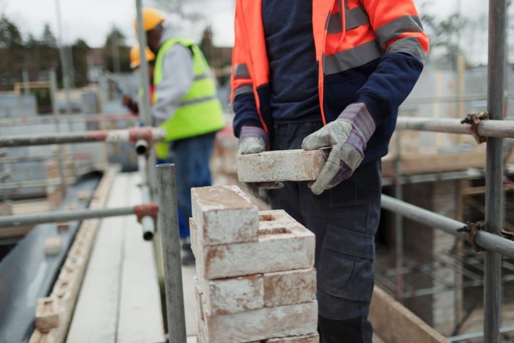 Construction worker in high-visibility clothing carefully placing bricks on a building site with scaffolding, emphasising safety and skill in the construction industry.