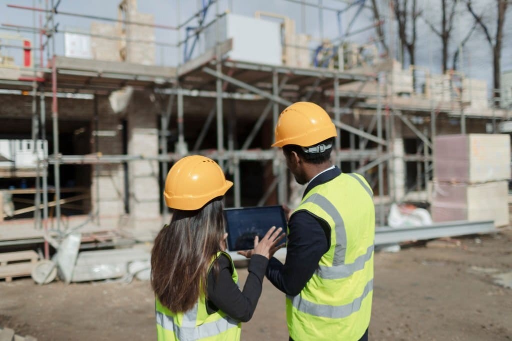 Two construction supervisors in safety gear discussing project details on a building site, with scaffolding and the structure of a house under construction in the background.