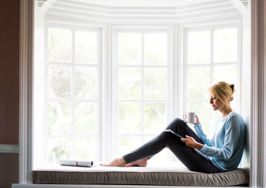 Serene image of a young woman enjoying her morning coffee while sitting on a cushioned window seat, reading a book in a cosy, well-lit nook with a bay window offering a view of greenery outside, embodying peaceful home living.
