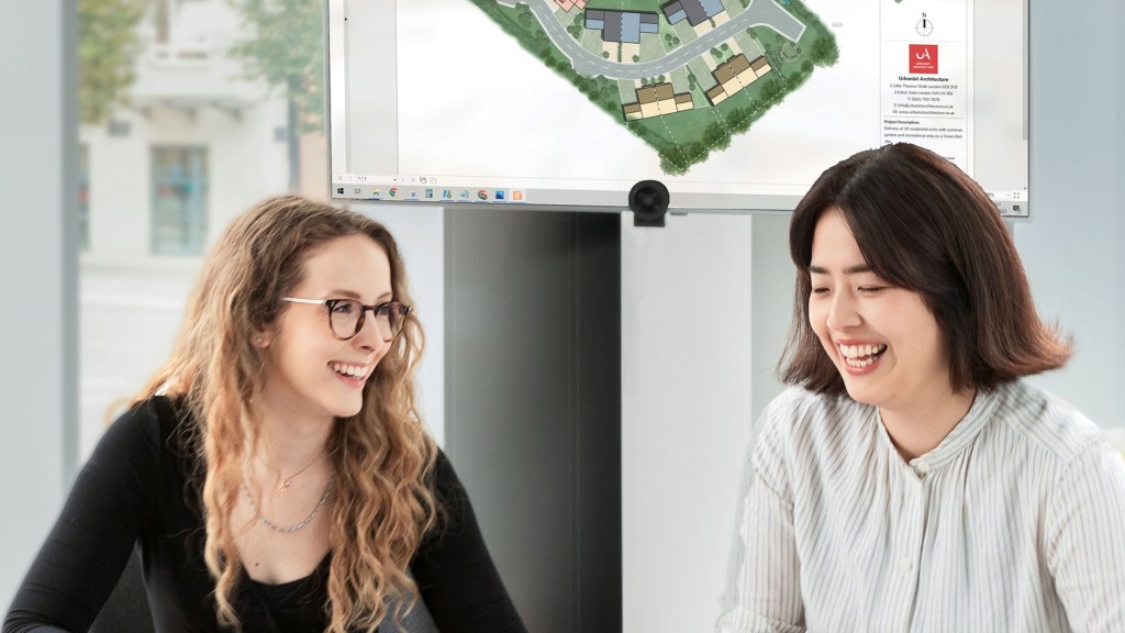 Two female colleagues laughing and sharing a joyful moment in front of a computer monitor displaying a 3D architectural site plan, illustrating a collaborative and positive work environment in a Greenwich, London, design studio.