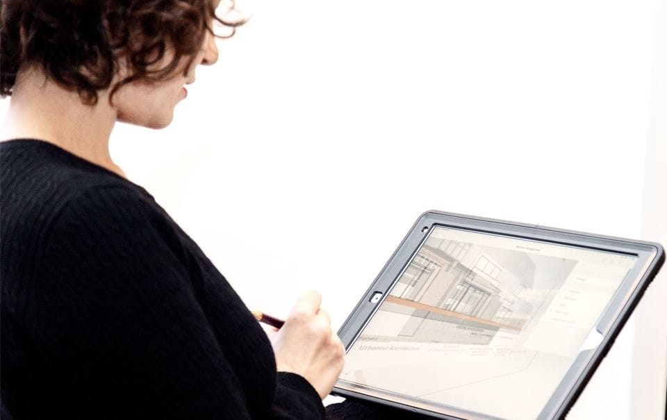 Focused professional reviewing architectural designs on a tablet, with a close-up on the digital screen showing detailed building plans, indicative of modern technology integration in architecture and design workflows.