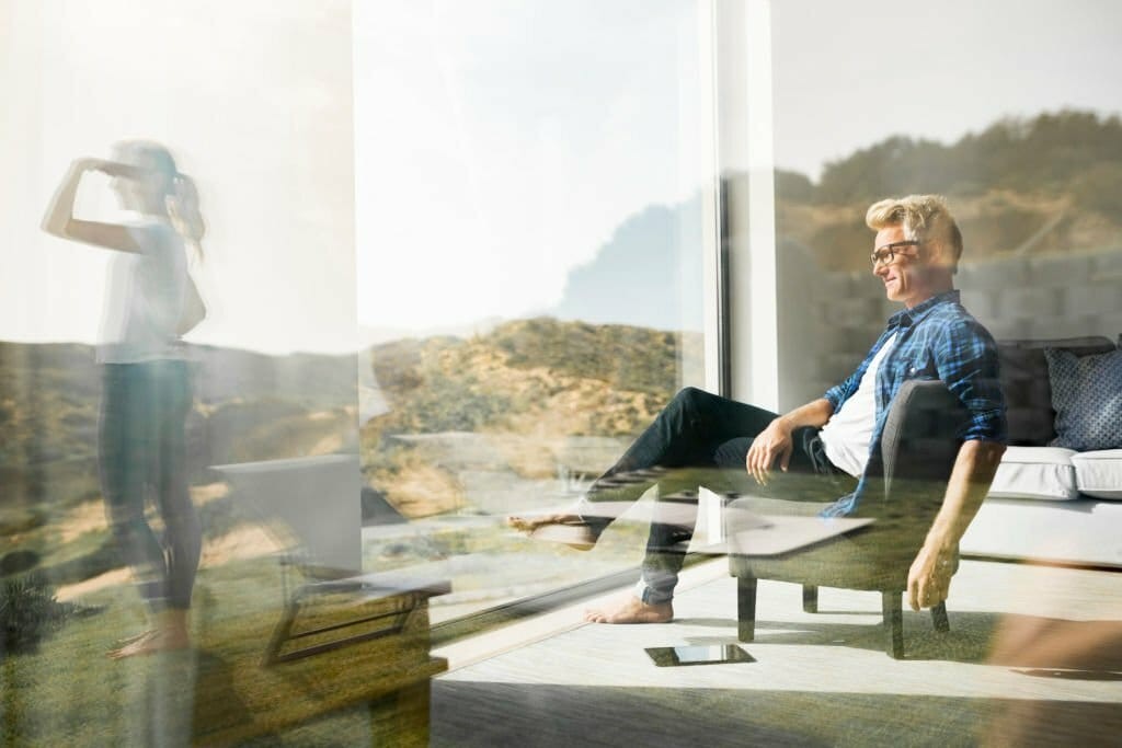 Relaxed man smiling and enjoying the view in a sunlit modern living room with floor-to-ceiling windows, while a woman stands outside on the patio, against a backdrop of a serene landscape, depicting contemporary home living.