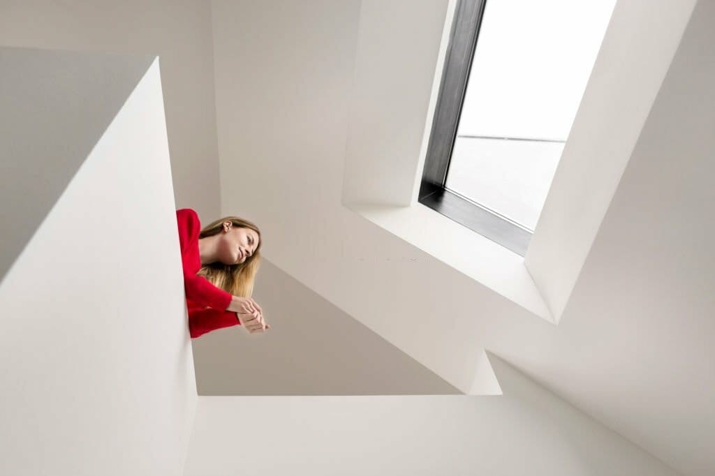 Upward perspective of a woman in a red sweater deep in thought, leaning on indoor balcony in a modern white interior with geometric shapes and a skylight window, emphasising contemporary architectural design and natural lighting.