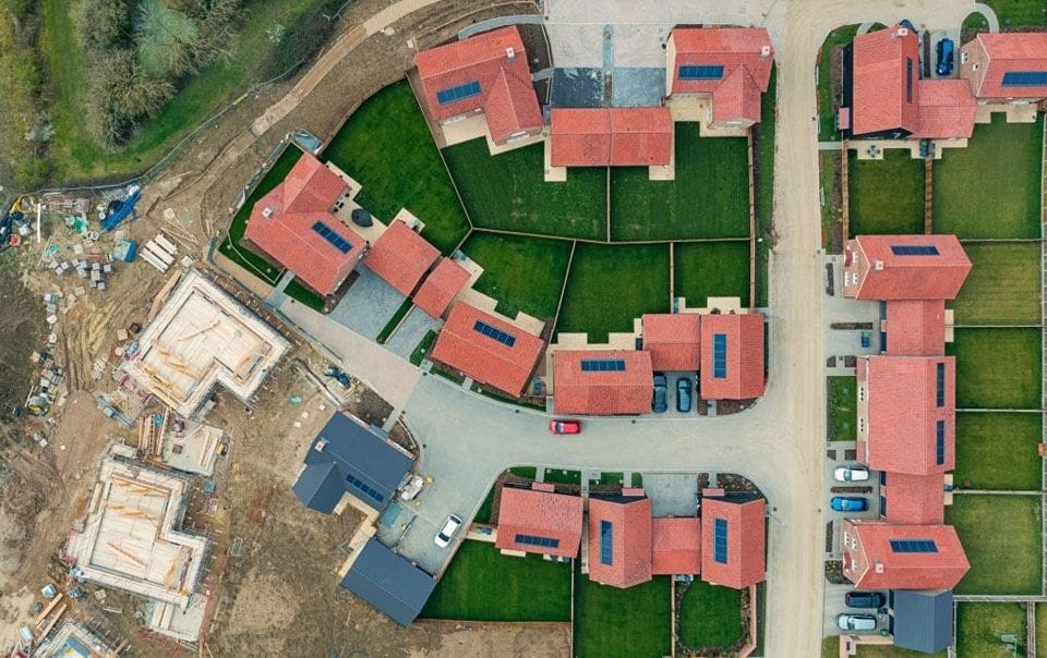An aerial view of a residential development on a small urban site, featuring modern homes with red roofs and solar panels, surrounded by green lawns and construction areas in progress.