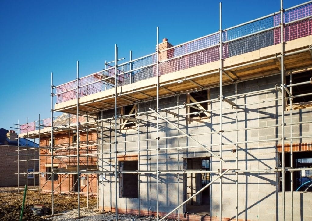 Construction site of a small residential development with scaffolding surrounding a partially built house, featuring exposed brickwork and concrete walls under clear blue skies.