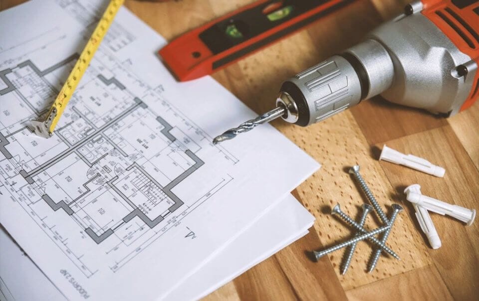 A complete set of construction drawings lay on a wooden table alongside an opened tap measure, level bubble ruler, cordless green power drill, plastic wall plugs and screws