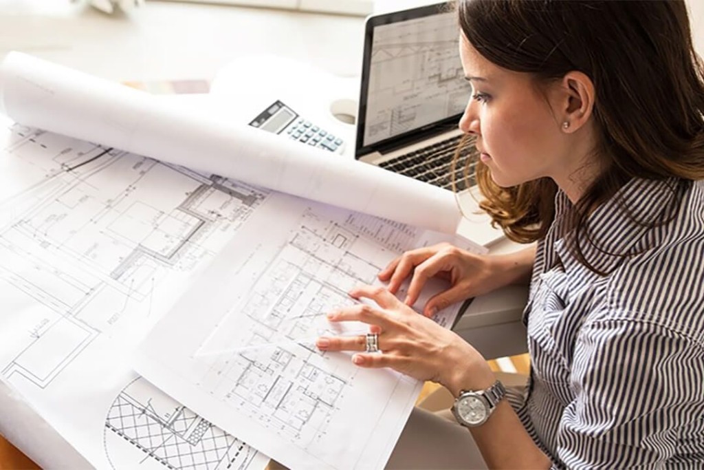 Female engineer in a black and white striped blouse conducting her design review process prior to construction with technical packages printed on various paper sizes and on her small laptop screen on the side
