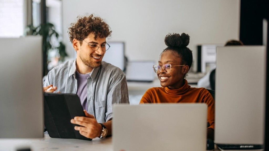 Two colleagues collaborating in a modern office environment; a man with curly hair holding a tablet and a woman with a bun smiling at the screen of her laptop.