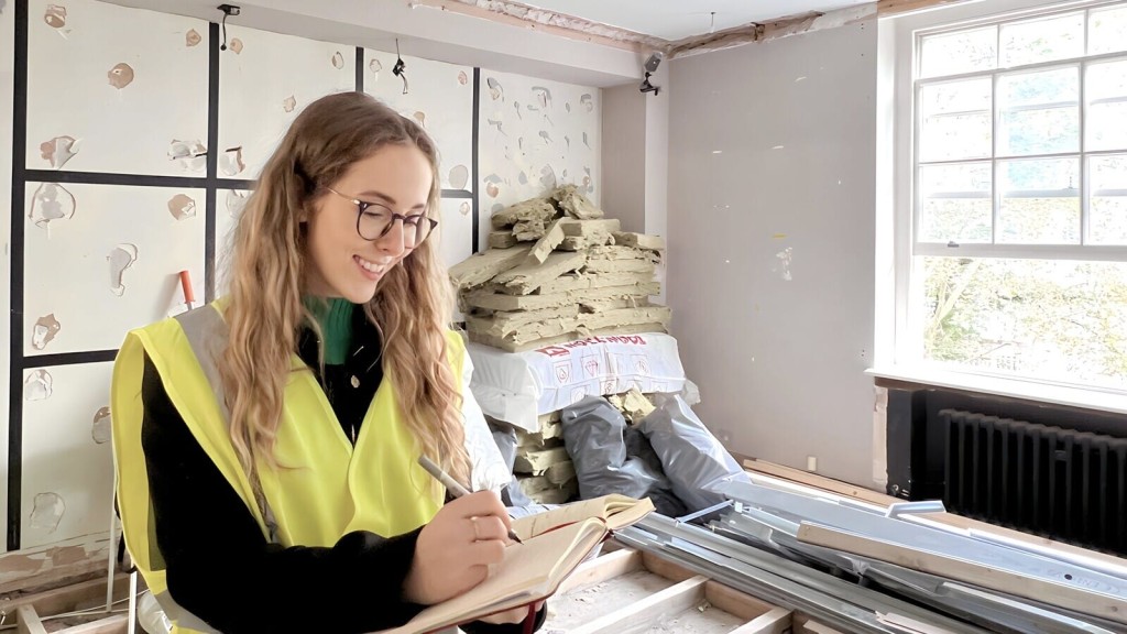 Smiling female construction supervisor in safety vest taking notes in a notebook in a room under renovation with exposed walls and windows.