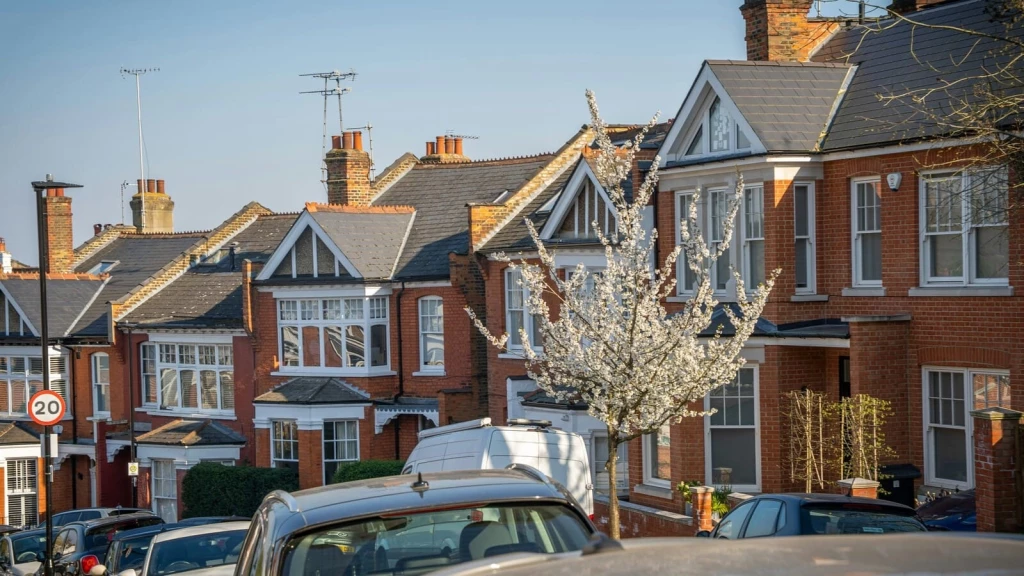 Row of red-brick terraced houses in London with bay windows, pitched roofs and cars parked along the street.