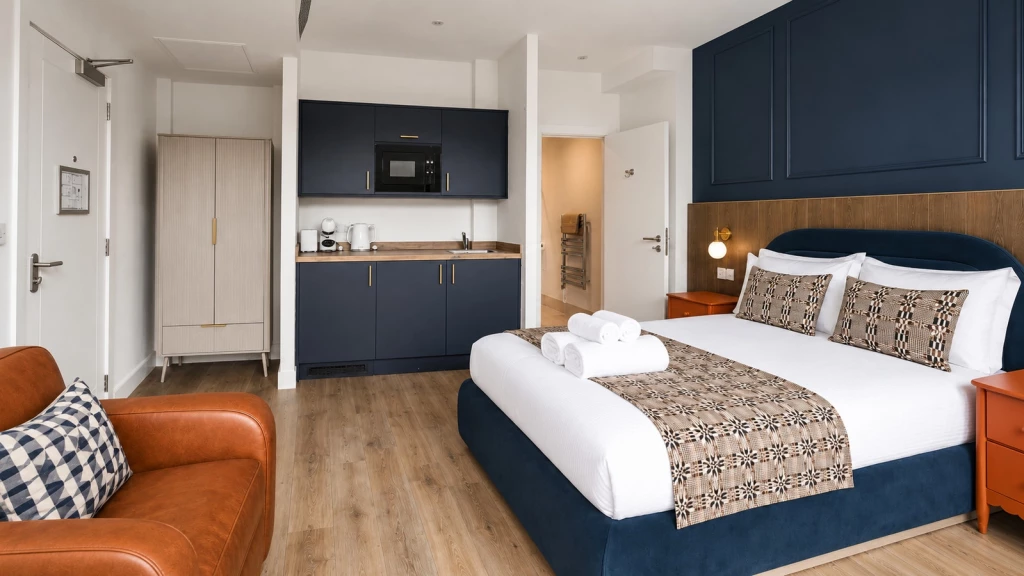 HMO room in London designed by Urbanist Architecture, with a double bed, en-suite bathroom, compact kitchenette with navy cabinetry, a wardrobe, an orange armchair and wood-effect flooring.