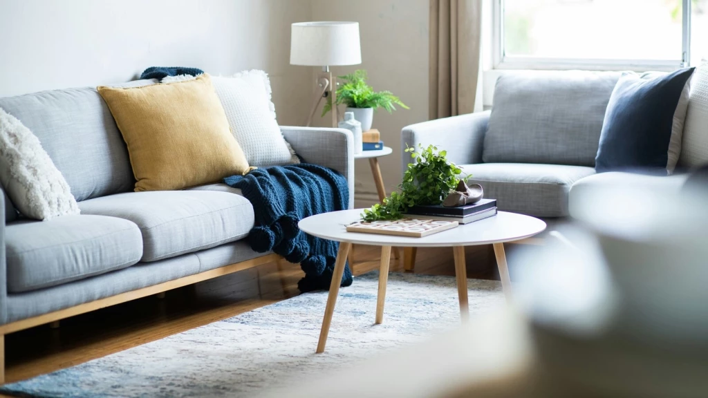 Shared living room in a London HMO property, with light grey sofas, mustard and navy cushions, a round coffee table, indoor plants and daylight from a large window.