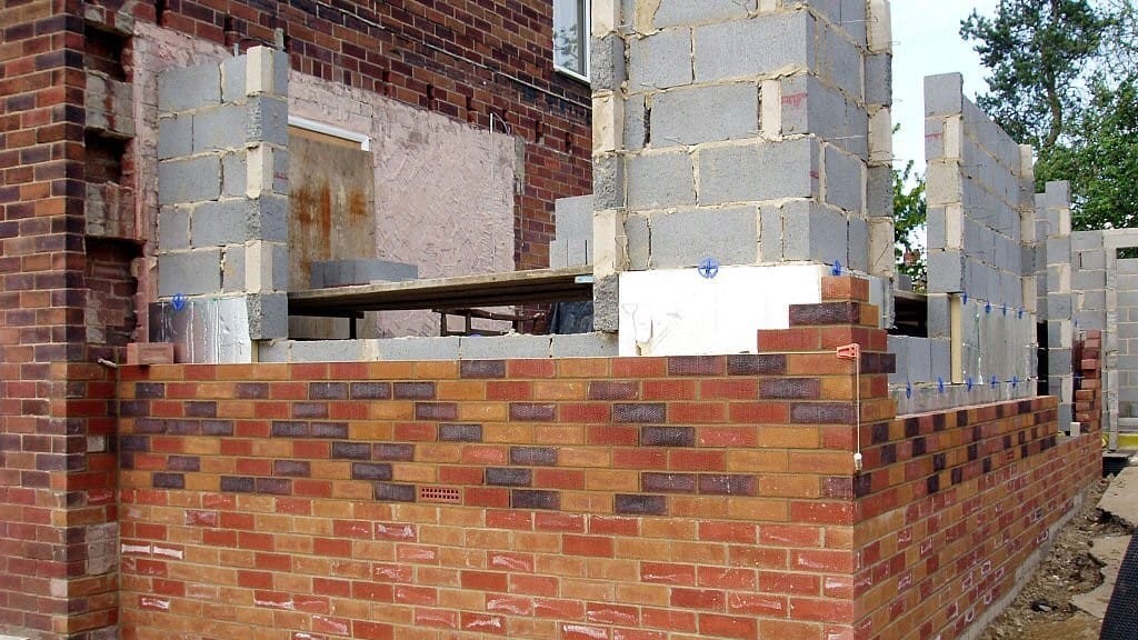 Construction in progress of a double-storey house extension with new brickwork walls rising from the foundations, integrating with the existing building structure, demonstrating the process of home renovation and expansion.