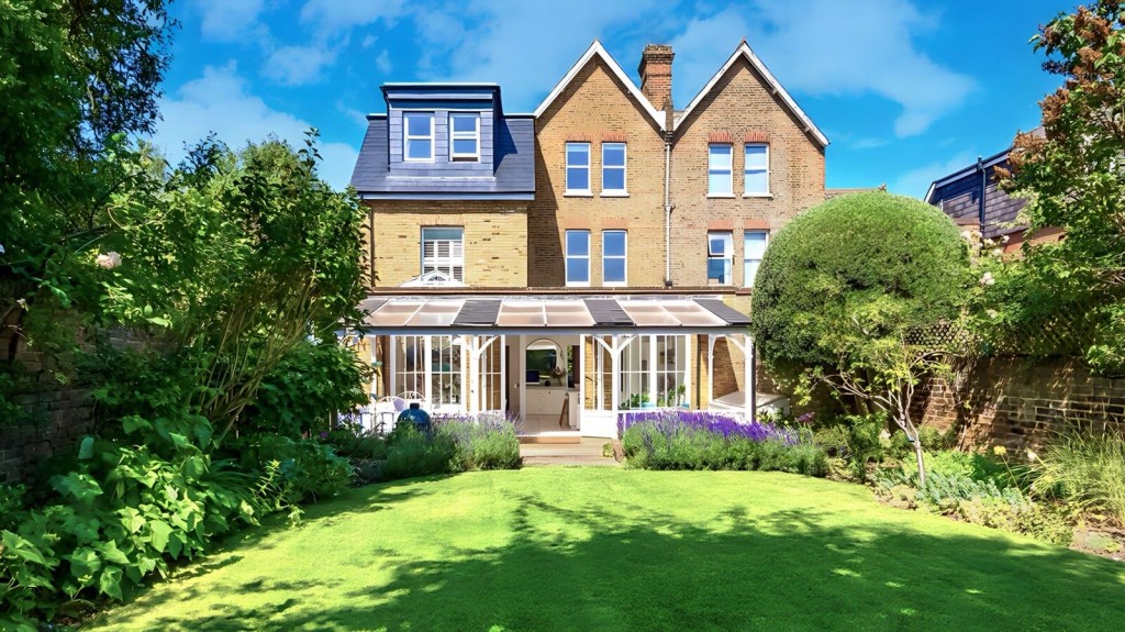 Charming Victorian-style tripe-storey house, featuring a glass conservatory, surrounded by a well-manicured garden with lush greenery and flowering plants, showcasing a classic architectural beauty.
