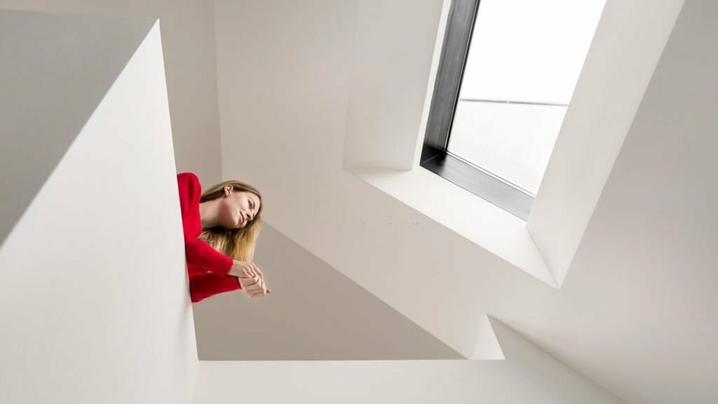 Woman in a red sweater relaxing in a minimalist attic space with white walls and angular lines, looking out of a large rectangular skylight.
