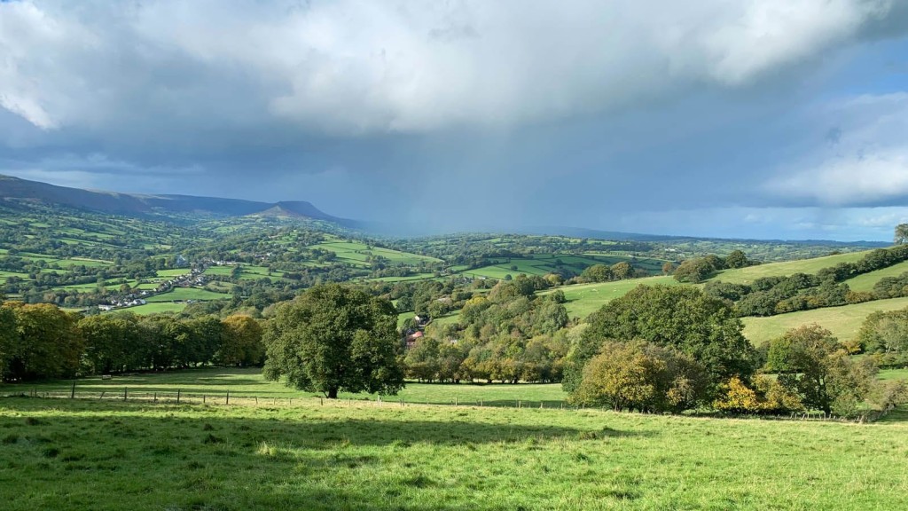 Panoramic view of a lush green countryside in England, featuring rolling hills, dense clusters of trees, and a distant village nestled in the valley. The dramatic sky, with a mix of clouds and sunlight, enhances the natural beauty of this rural landscape, exemplifying the types of areas where eco-house planning permission challenges might arise.