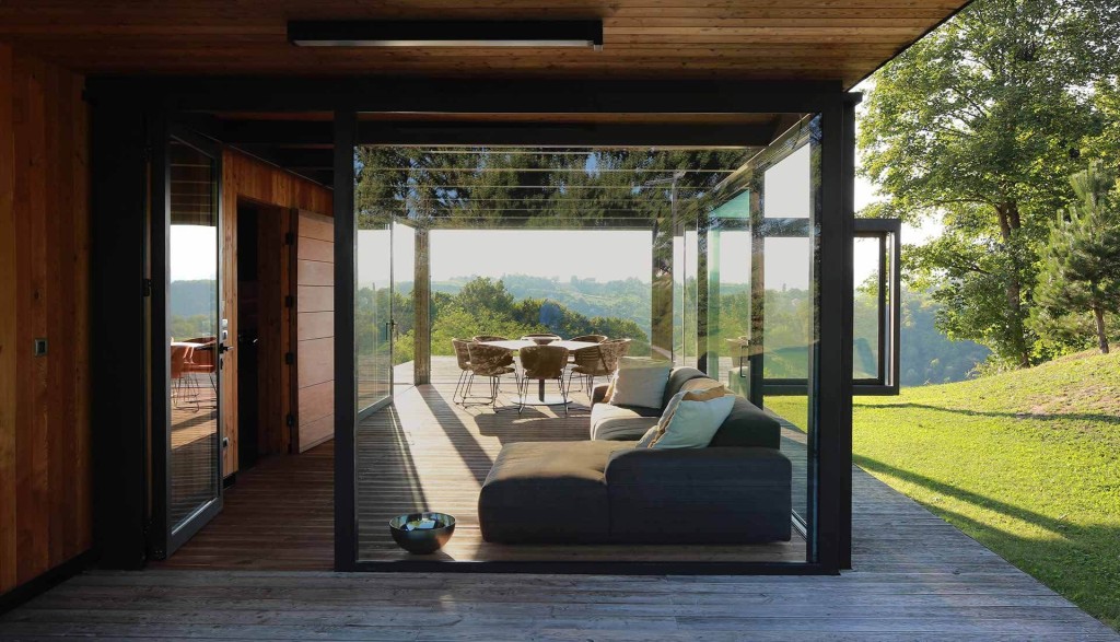 Modern eco-house with large glass doors opening to a wooden deck, showcasing an outdoor dining area with scenic views of lush greenery and distant hills. The sustainable home features natural wood elements and open spaces that blend seamlessly with the environment.