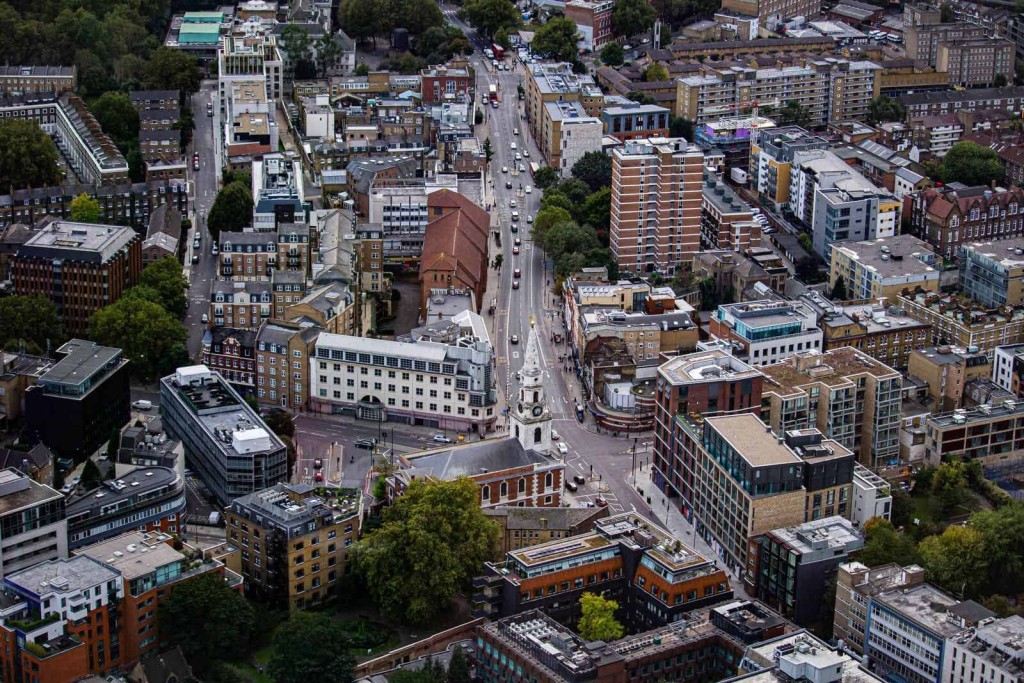 Bird's-eye view of an urban street intersection with a variety of residential and commercial buildings, showcasing diverse architectural styles and bustling city life, with visible cars and greenery interspersed among the structures.