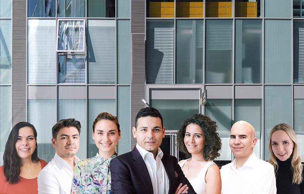 Team of seven professional architects with a diverse mix of men and women, smiling confidently in front of a modern office building facade with reflective glass windows, representing the collaborative spirit of contemporary architectural design.