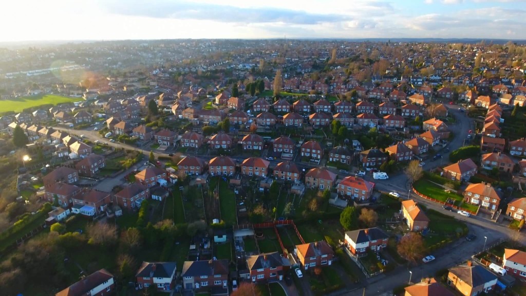 An aerial view of a sprawling suburban neighbourhood featuring numerous semi-detached and detached houses with red-tiled roofs. The layout is organised with curving streets and lush green gardens, giving a sense of spaciousness and community. The image highlights the potential for upward extensions in suburban homes, providing additional living space without expanding the building footprint. This picturesque neighbourhood exemplifies the type of residential area where extending houses upwards could enhance property value and living conditions.